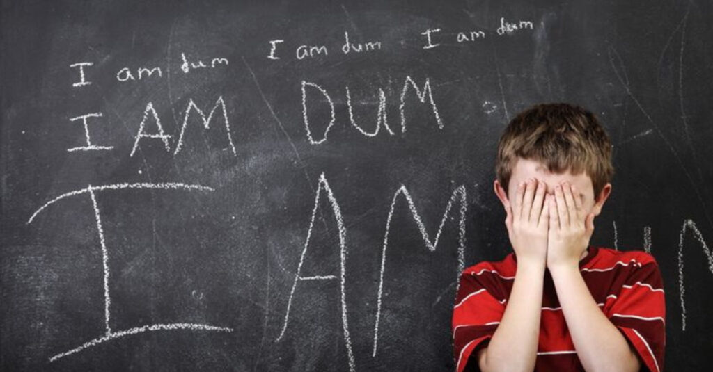 "A child stands in front of a chalkboard, their hands covering their face in a gesture of grief and frustration. The chalkboard behind them is filled with the words 'I am dum' written repeatedly, illustrating a poignant moment of self-doubt and emotional distress."